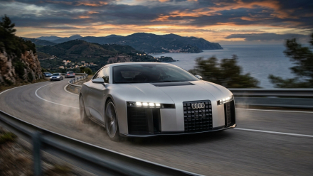 Audi Nuvolari Concept Speeds Silver Audi Nuvolari performance concept driving on a winding coastal mountain road at sunset, with headlights illuminated and the sea visible in the background.