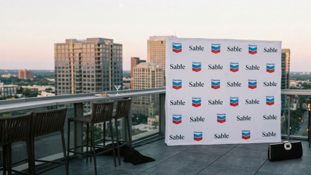 Rooftop event setting overlooking a city skyline, featuring a branded backdrop with Chevron logos and Sable Offshore names, bar stools, cocktail glass, and a black handbag in the foreground.