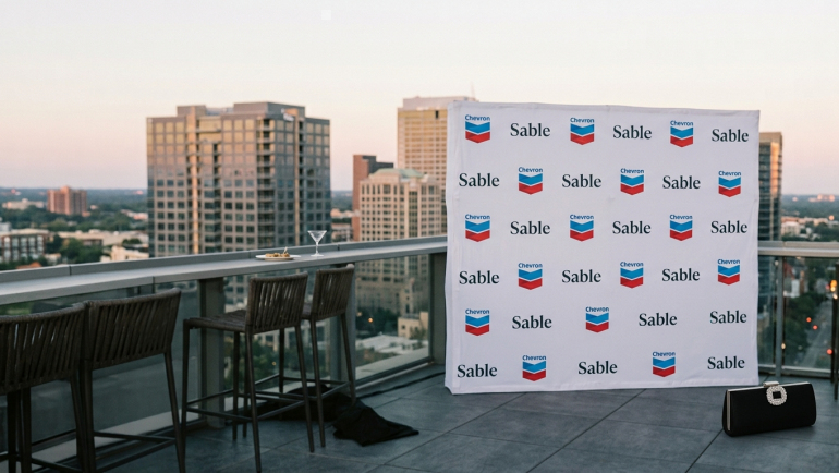 Rooftop event setting overlooking a city skyline, featuring a branded backdrop with Chevron logos and Sable Offshore names, bar stools, cocktail glass, and a black handbag in the foreground.